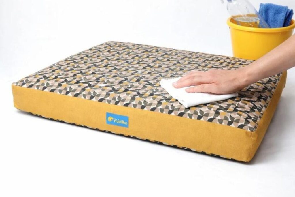 Uma mão humana passa um pano branco dobrado sobre a superfície de um colchão pet retangular plano com estampa geométrica cinza e amarela e laterais amarelas. No canto superior direito, um balde amarelo contém um frasco borrifador e um pano azul, tudo sobre um fundo branco.
