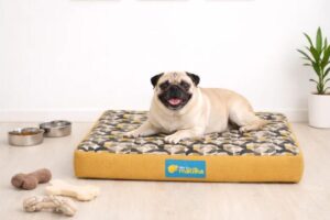 Um cachorro da raça Pug deitado de forma confortável e sorridente sobre uma caminha retangular amarela com estampa geométrica, em um ambiente com piso claro e parede branca.