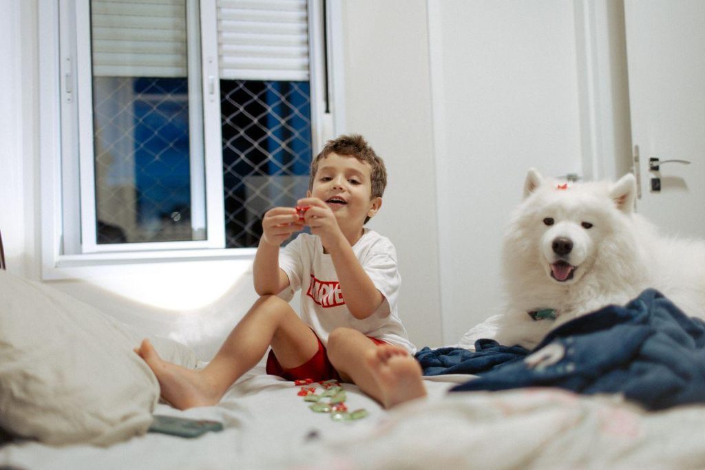 Criança sentada na cama ao lado de um cachorro branco em um quarto residencial.